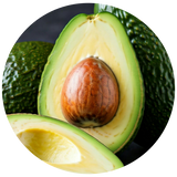 A close-up circular photo of an avocado cut open, showing the green flesh, creamy yellow interior, and large brown seed, with additional whole avocados in the background.