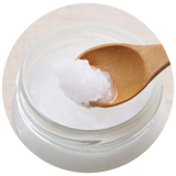 A close-up of a clear glass jar filled with a white organic coconut oil, with a wooden spoon lifting a scoop of the product against a light neutral background.