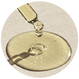 A close-up circular shot of a clear liquid droplet dripping from a glass pipette onto a glossy pool of transparent beige-tinted gel or oil on a light textured surface.